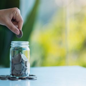 Closeup image of a hand collecting and putting coins in a glass jar for saving money concept